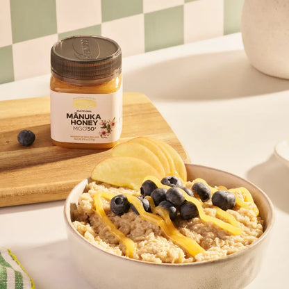 Bowl of oatmeal with honey and blueberries next to a jar of Manuka Honey on a wooden board.