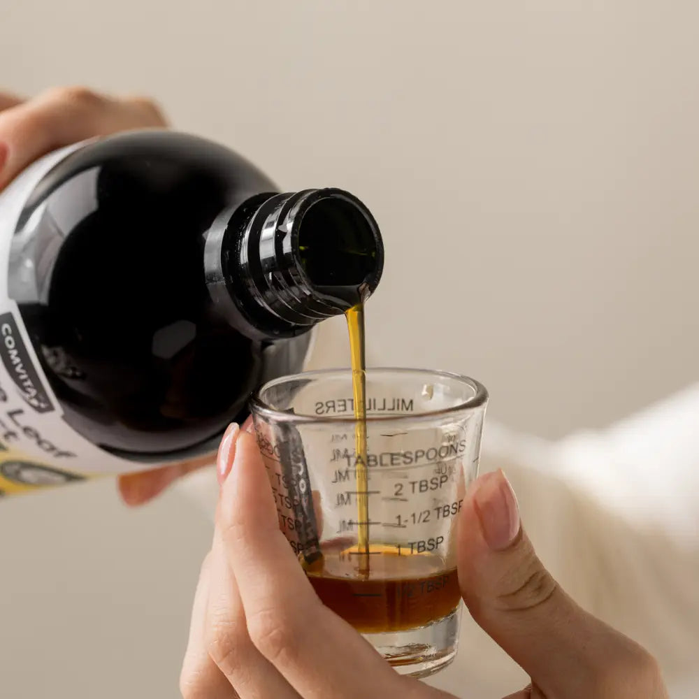 Person pouring Olive Leaf Extract from a bottle into a measuring cup with a neutral background