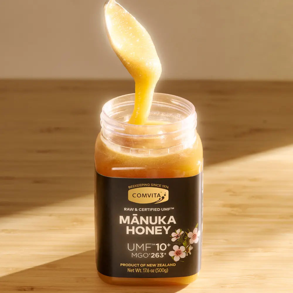 Jar of Comvita Manuka Honey with a spoonful being poured on a wooden surface