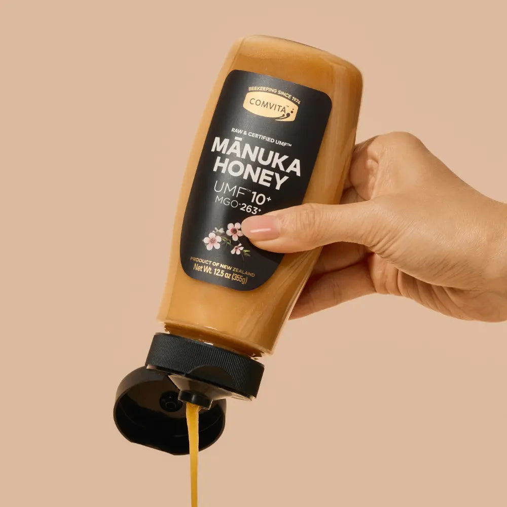 Hand holding a bottle of Comvita Manuka Honey with visible honey flow.