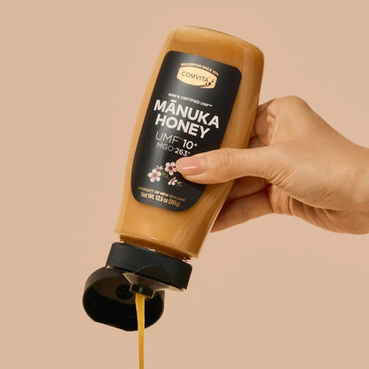 Hand holding a bottle of Comvita Manuka Honey with visible honey flow.