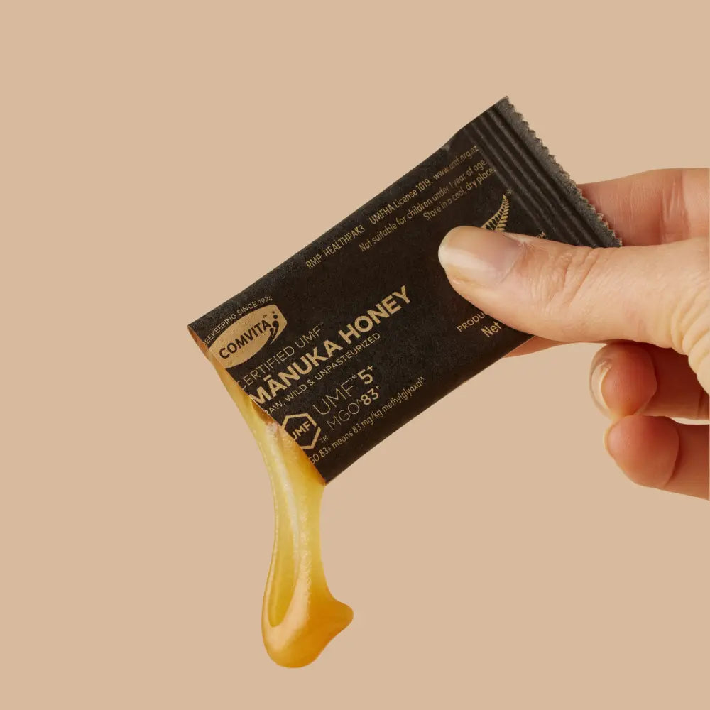 Manuka honey single tear pack with visible honey dripping, held by a hand against a beige background.
