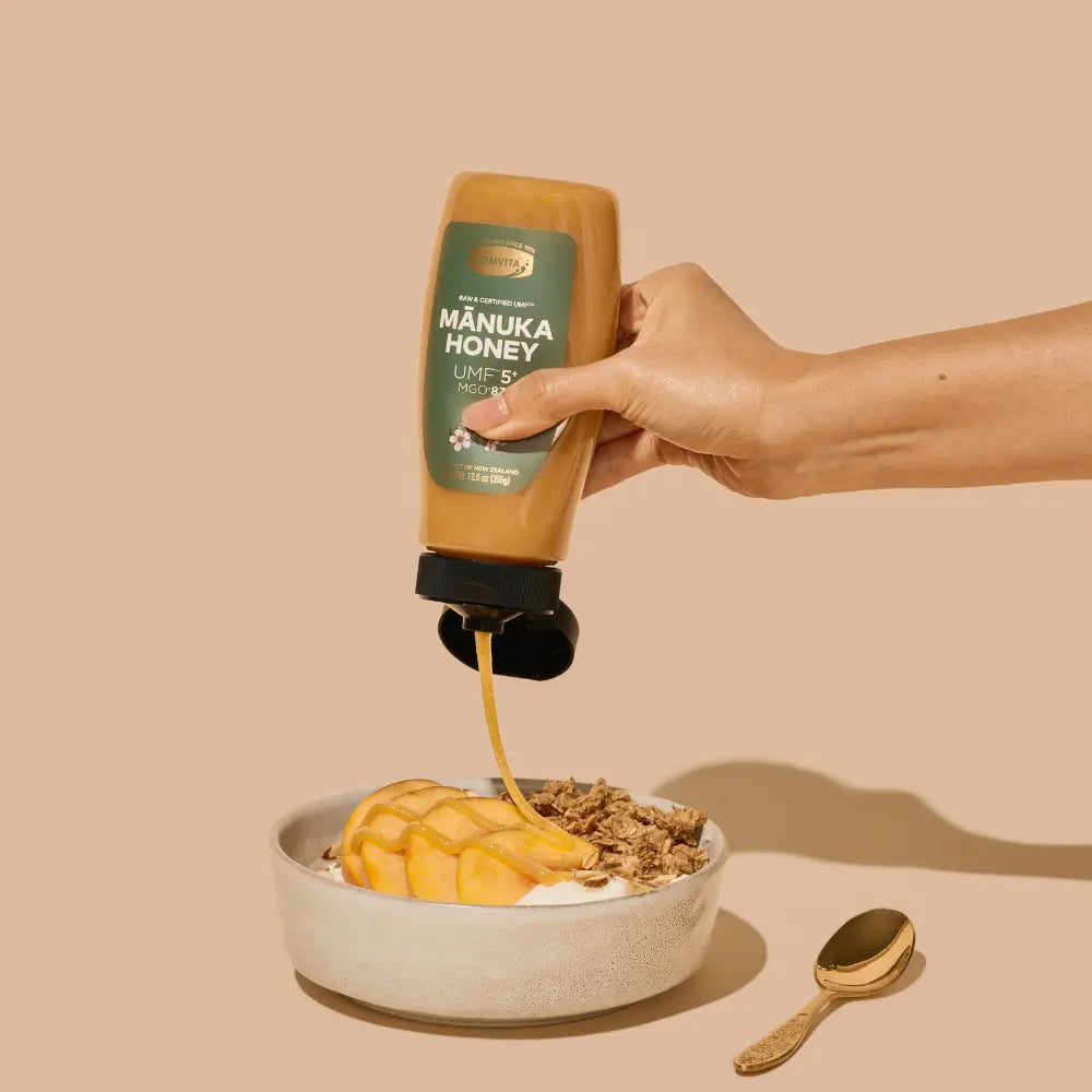 Hand pouring Manuka Honey into a bowl of yogurt and granola on a beige background